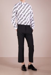 Witte overhemd met knopen en een zwart diagonaal tekstpatroon met de tekst "VERSUS," gecombineerd met zwarte op maat gemaakte broek en glanzende zwarte schoenen.
