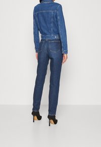 Chaqueta de mezclilla azul oscuro y vaqueros a juego, ambos con un diseño ajustado, costuras visibles y bolsillos traseros clásicos. Zapatos de tacón alto negros.