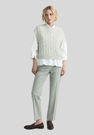 Woman wearing light gray cable knit sleeveless sweater over white long-sleeve shirt, gray pinstripe pants, and dark brown loafers, standing.
