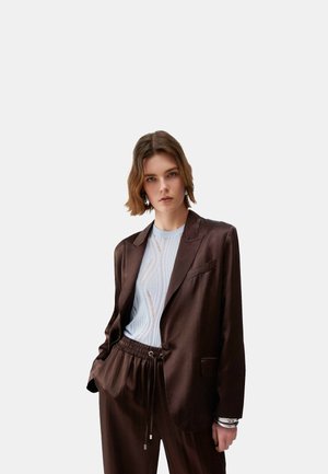 Giovane donna che indossa un blazer marrone scuro e pantaloni abbinati sopra una maglia bianca strutturata, in piedi con una mano in tasca contro uno sfondo semplice.