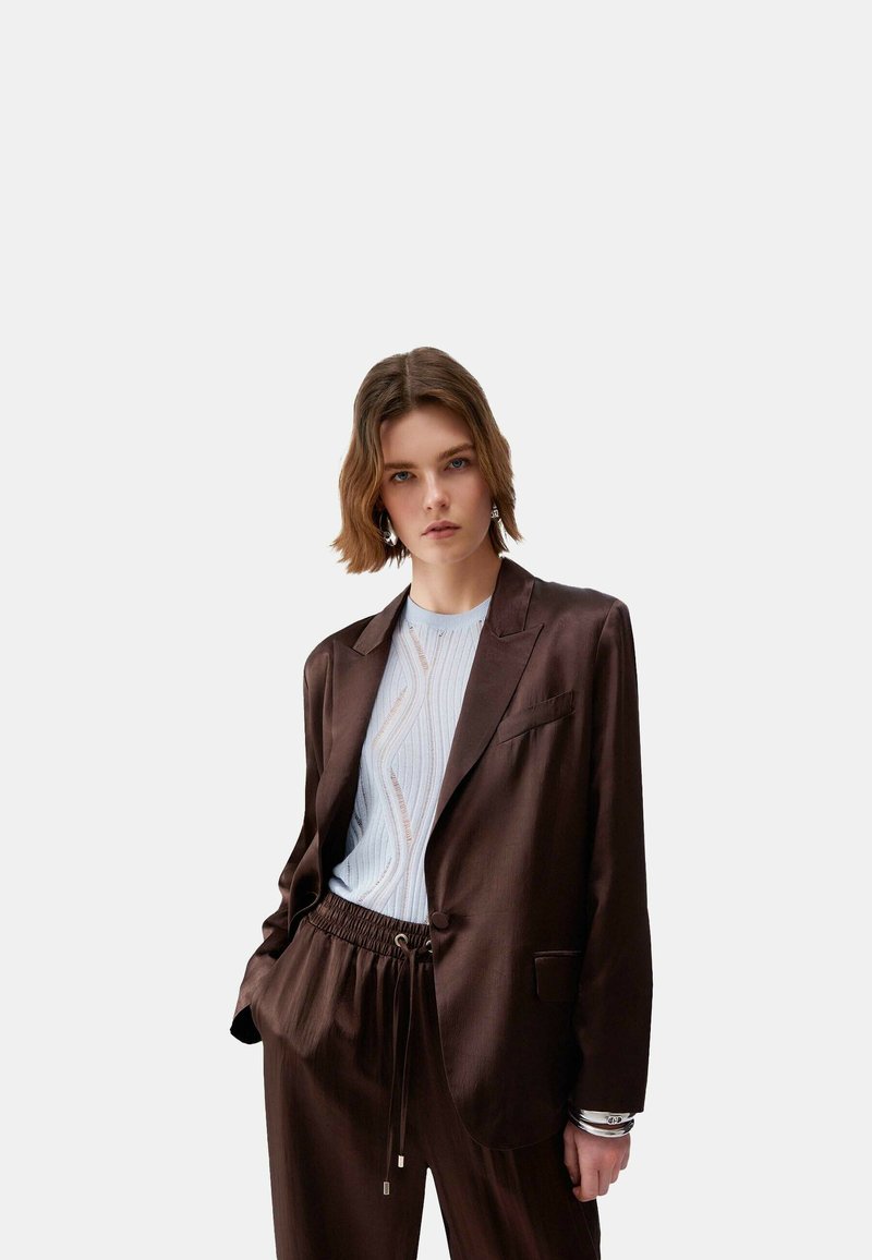 Mladá žena v tmavohnedom saku a zodpovedajúcich nohaviciach cez biely textúrovaný top, stojaca s jednou rukou v vrecku pred jednofarebným pozadím.