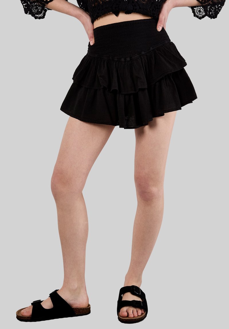 Falda negra con volantes en capas y cintura fruncida, hecha de una tela ligera. Combinada con sandalias negras que cuentan con dos correas ajustables.