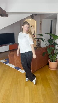 Valkoinen t-paita, jossa pieni musta logo, mustat housut hopeanvärisillä pilkuilla ja hopeiset kengät puulattialla, taustalla sisäkasveja.