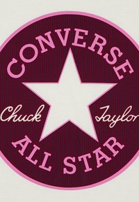 Converse Chuck Taylor All Star-logo med en hvit stjerne på en burgunder bakgrunn, kantet med rosa tekst og fin ribbet tekstur.