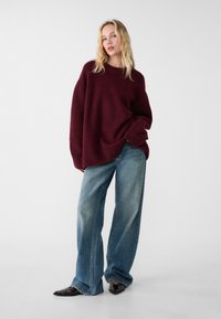 Pull en maille oversize bordeaux avec un col rond, associé à un jean large bleu clair et des chaussures à bouts pointus noires ornées.