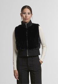 Čierny umelejší kožuch s vysokým golierom, zapínaním na zips a rebrovanou tmavou látkou na leme. Model má pod ním dlhý rukáv.