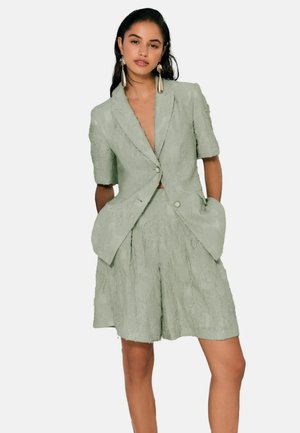 Frau trägt einen strukturierten, salbeigrünen Kurzarm-Blazer und passende kniehohe Shorts, accessoriert mit langen metallischen Ohrringen.