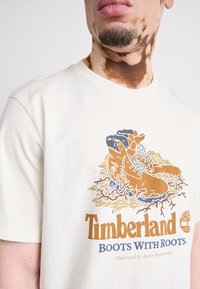 Kremasta pamučna majica s ilustracijom čizama s korijenjem i natpisom "Timberland ČIZME S KORIJENJEM" u smeđoj i plavoj boji.