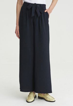 Frau trägt schwarze, weit geschnittene Hose mit hohem Bund, mit einem Stoffgürtel, der zu einer Schleife gebunden ist, und goldenen Slip-On-Loafern.