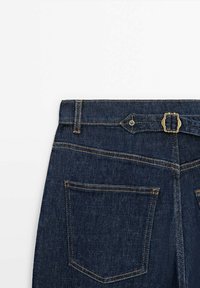 Dunkelblaue Jeanshose von hinten gezeigt, mit Gürtelschlaufe, einem kleinen verstellbaren Riemen mit einer Metall-Schnalle und zwei genähten Gesäßtaschen.