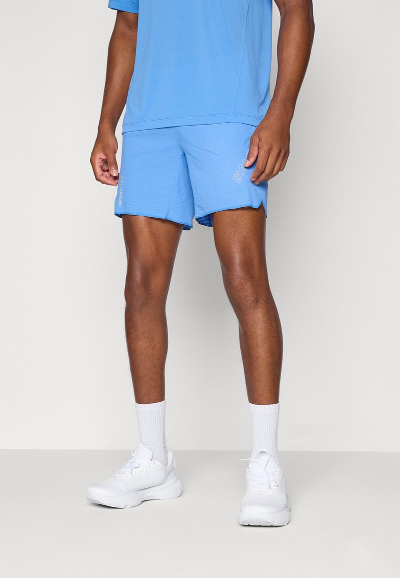 Pantalones cortos deportivos azules con una tela texturizada, aberturas laterales y un detalle de logo, combinados con calcetas blancas y zapatillas deportivas.