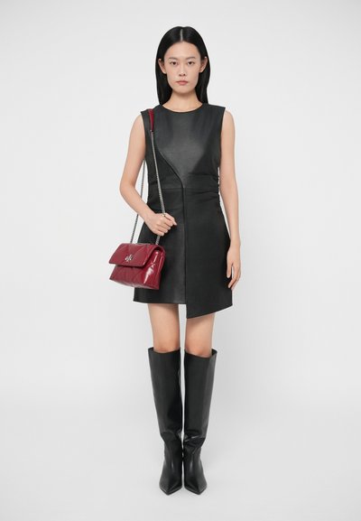 Μαύρο δερμάτινο φόρεμα χωρίς μανίκια με ασύμμετρο σχέδιο, συνδυασμένο με μαύρες μπότες ως το γόνατο και γυαλιστερή μπορντό τσάντα χειρός με ασημένια αλυσίδα.