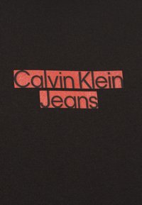 Tela negra con bloques rectangulares rojos que contienen el logo de "Calvin Klein Jeans" en texto negro centrado sobre el material.