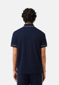 Mörkblå pikétröja med vit kant, korta ärmar och klassisk krage. Mjukt tyg med avslappnad passform, visas bakifrån.