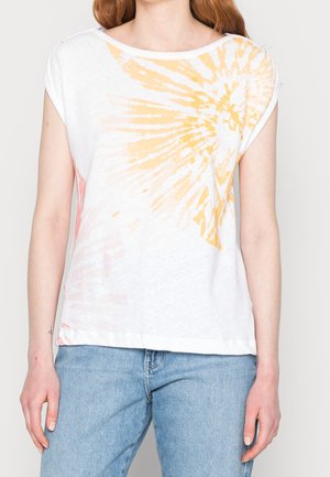 Person iført en hvid kortærmet bluse med gul batikmønster og blå denimjeans, stående mod en ensfarvet baggrund.