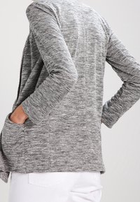 Šedý melírovaný cardigan s dlouhými rukávy, otevřeným předním dílem a bočními kapsami. Měkká, texturovaná tkanina s volným střihem a jemnými detaily švů.