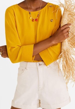 Vrouw draagt een mosterdgele top met lange mouwen en knopen, witte shorts met een hoge taille, kleurrijke kralenketting en houdt een strohoed met franjes vast.