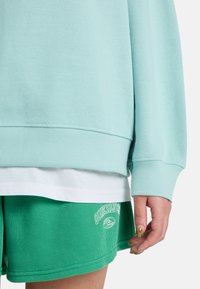 Φλις φούτερ σε μέντα με ριμπ μανσέτες, φορεμένο πάνω από άσπρη μπλούζα, συνδυασμένο με πράσινα βερμούδα από κοτλέ με κέντημα λογότυπου.