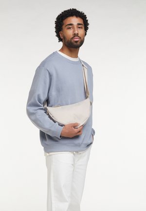 Hombre con cabello rizado que lleva una sudadera azul y pantalones blancos, sosteniendo un bolso bandolera beige sobre un fondo blanco liso.