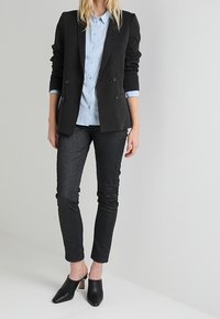 Blazer noir à double boutonnage sur une chemise à boutons bleu clair, associé à un pantalon slim-texturé foncé et des mules noires à talon bloc.