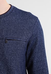 Maglione a maglia blu con texture, dotato di tasca frontale con zip orizzontale e scollatura rotonda, indossato da una persona su uno sfondo semplice.