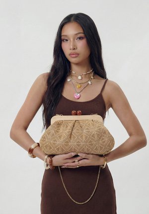 Jonge vrouw in een bruine jurk houdt een gestructureerde beige clutch vast, draagt gelaagde kettingen en armbanden van gemengde materialen tegen een effen achtergrond.