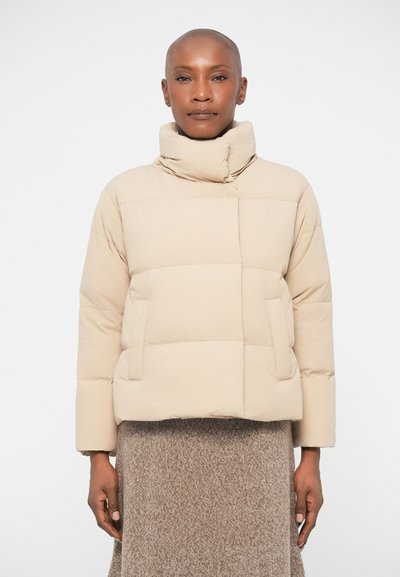 Doudoune beige avec un col haut, design matelassé et poches avant. Tissu texturé avec une finition lisse. Portée sur une jupe en tricot marron.