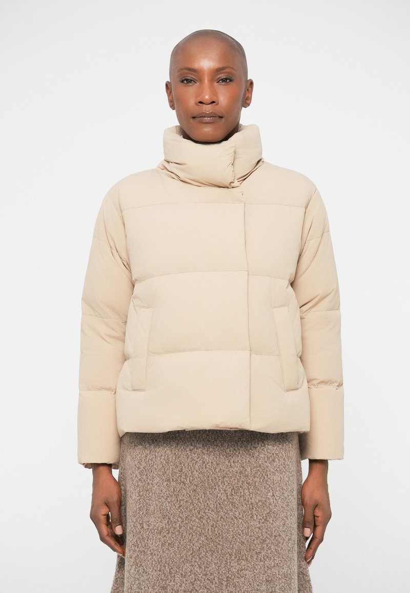 Beige puffer jacket with a high collar, quilted design, and front pockets. Textured fabric with a smooth finish. Worn over a brown knit skirt.