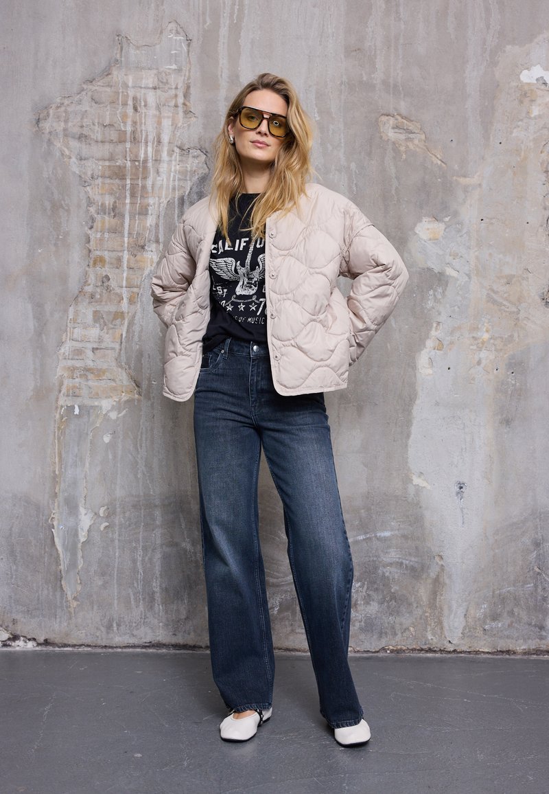 Femeie purtând o geacă matlasată bej supradimensionată, tricou negru cu imprimeu grafic, blugi din denim închis la culoare, pantofi albi și ochelari de soare cu lentile galbene, stând sprijinită de un perete texturat.