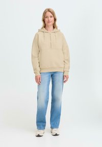 Beige hooded sweatshirt with front kangaroo pocket paired with light blue straight-leg jeans and light-colored sneakers. Plain background.