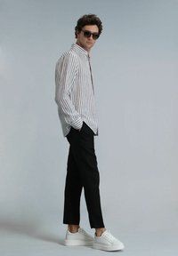 Chemise à rayures blanche et marron, pantalon noir et baskets blanches. Le modèle porte des lunettes de soleil, avec les cheveux coiffés en boucles lâches.