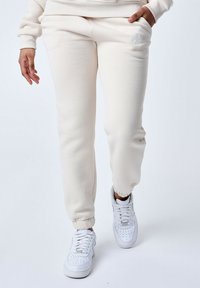 Pantalones de chándal beige con puños elásticos, con bolsillos laterales y un logo bordado en blanco. Llevados con zapatillas blancas sobre un fondo neutro.