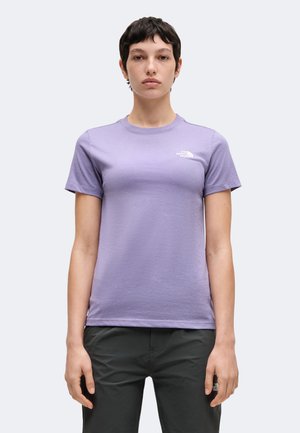 Persoon met kort donker haar die een lichtpaarse The North Face T-shirt en een donkere broek draagt, staand tegen een effen lichte achtergrond.