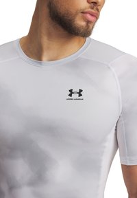 Witte getailleerde sportshirt met korte mouwen, gladde textuur en een zwart Under Armour-logo op de borst.