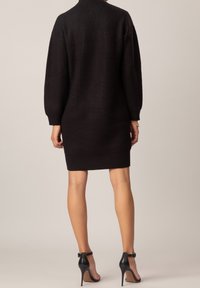 Robe noire en maille avec une coupe décontractée, texture côtelée et manches longues, arrivant au-dessus du milieu de la cuisse. Associée à des talons noirs à bride cheville.