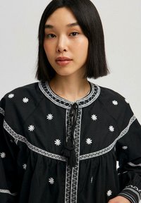 Black embroidered blouse featuring white floral patterns, contrasting stitching, a round neckline, and a tassel tie closure.