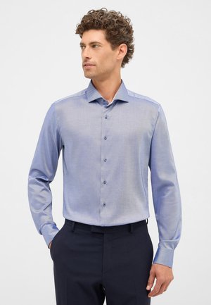 Chemise boutonnée bleu clair avec une texture lisse, présentant un motif subtil, un col écarté et des boutons assortis. Portée avec un pantalon sombre.