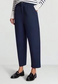 Pantaloni navy a vita alta e gamba larga con pieghe frontali, tasche laterali e un bottone dorato. Abbinati a mocassini neri lucidi.