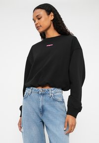 Sweatshirt negru cu logo roz, cu mâneci lungi și croi lejer. Asortat cu blugi de culoare albastră deschisă, cu talie înaltă, având un detaliu distrus.