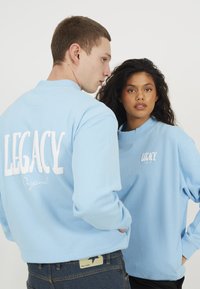 Lyseblå bomullssweatshirts med hvit tekst "LEGACY" og "Original" trykt på rygg og front, med ribbestrikkede krager og mansjetter.
