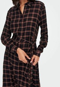 Frau in einem langärmeligen schwarzen Kleid mit rotem und weißem Karomuster, mit durchgehender Knopfleiste und gebundenem Taillengürtel, der den Stoff anpasst.