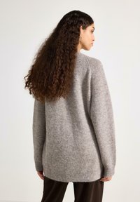 Oversize striktrøje i gråmeleret farve med lange ærmer, høj/lav kant og blød tekstur, kombineret med mørkebrune bukser.