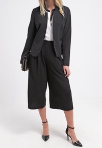Blazer noir ajusté sans revers, associé à des culottes larges noires. Le mannequin porte un chemisier blanc et des talons aiguilles noirs.