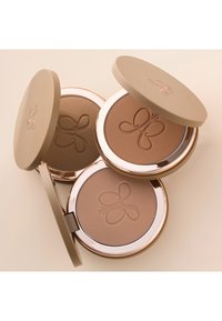 Drie ronde bronzerdozen met beige deksels en gouden accenten, met geëmbosseerde vlinderontwerpen op matte poeders in verschillende tinten.