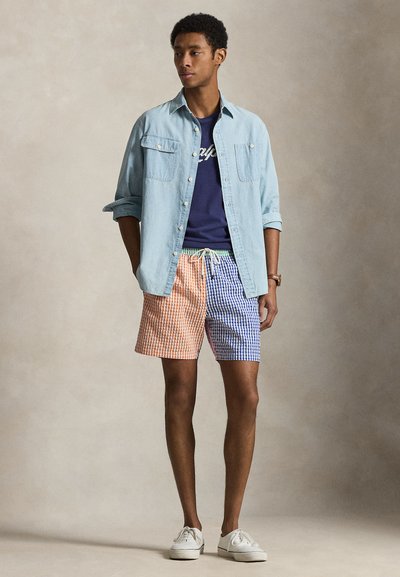 Άνδρας που φοράει ένα ανοιχτό μπλε τζιν πουκάμισο πάνω από ένα σκούρο μπλε μπλουζάκι με σχέδιο, πορτοκαλί και μπλε σορτς με καρό σχέδιο και άσπρα αθλητικά παπούτσια, στέκεται μπροστά σε απλό φόντο.