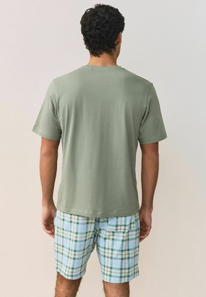 Mann mit lockigem dunkelbraunem Haar, der ein einfaches olivgrünes T-Shirt und blau-grün karierte Shorts trägt, steht mit dem Rücken zur Kamera.