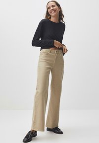 Beige, hochgeschnittene, ausgestellte Jeans mit Fronttaschen und glatter Textur, kombiniert mit einem enganliegendem schwarzen Langarmoberteil und schwarzen Schuhen.