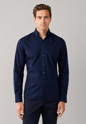 Jonge man met kort bruin haar die een nauwsluitend donkerblauw overhemd met knopen en een donkere broek draagt, staand tegen een effen achtergrond.