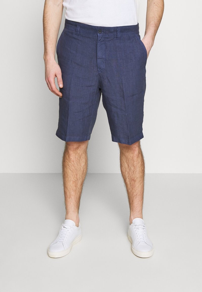 Homme portant un short bleu marine jusqu'aux genoux, des baskets blanches et un t-shirt blanc rentré dans le short, debout les mains dans les poches.