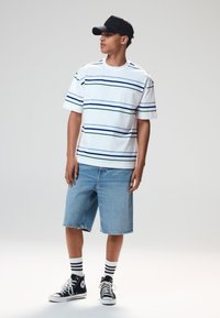 Maglietta bianca a maniche corte con righe orizzontali blu e verdi, abbinata a pantaloncini di jeans azzurri. Cappellino nero e sneakers alte.